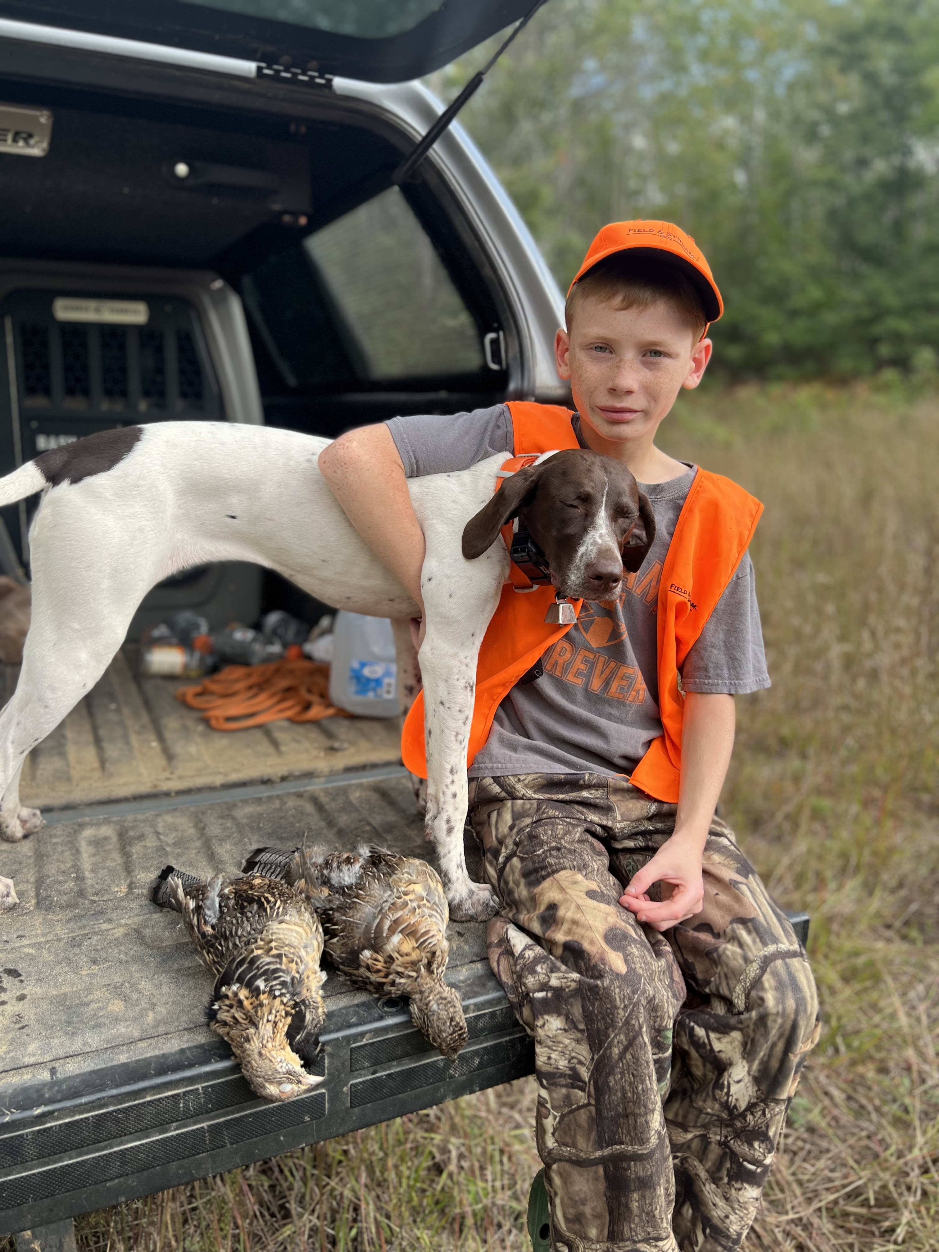 A boy hugs a German Shorthaired Pointer on a tailgate with two harvested grouse.