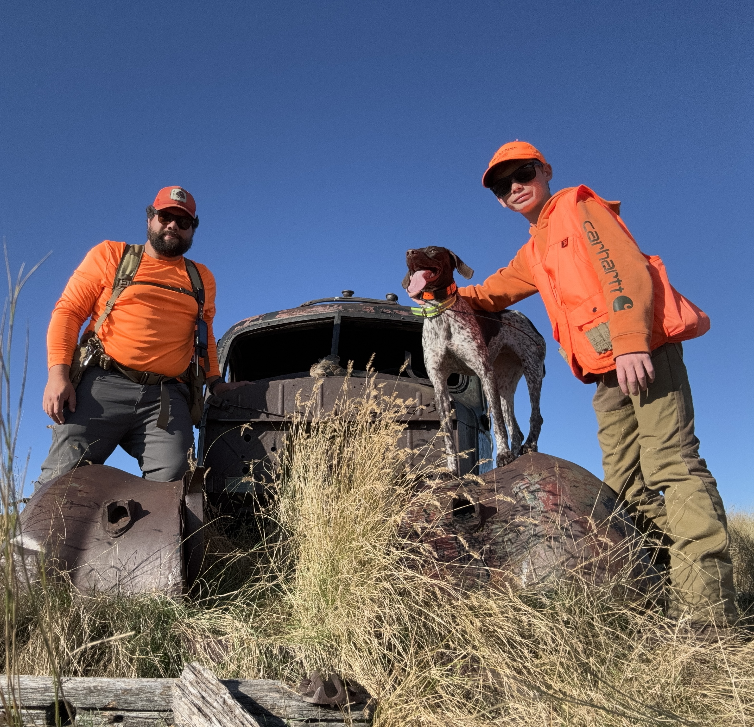 Two hunters and a German Shorthaired Pointer pose on an old rusted truck on the western prairie.